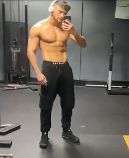 A shirtless man with headphones taking a mirror selfie in a gym.