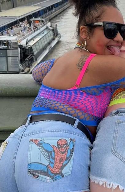 A woman wearing sunglasses, a bright pink bra, and a blue fishnet top, with denim shorts featuring a Spider-Man design on the back pocket, looks over her shoulder at the camera with her tongue out, with a river and boat in the background.
