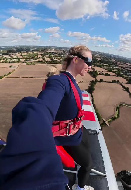 Sydney Sweeney wing walking, high above fields, secured in red harness and goggles.