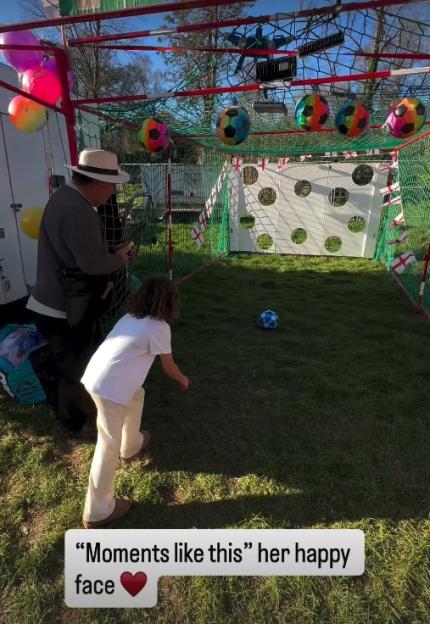 Man in a hat standing next to a child playing with a soccer ball in a grassy outdoor area with a soccer goal in the background.