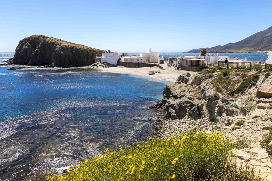 Coastal landscape of Isleta del Moro, Cabo de Gata Natural Park, with white houses on a sandy beach next to blue water, and a rocky hillside covered in yellow flowers in the foreground.
