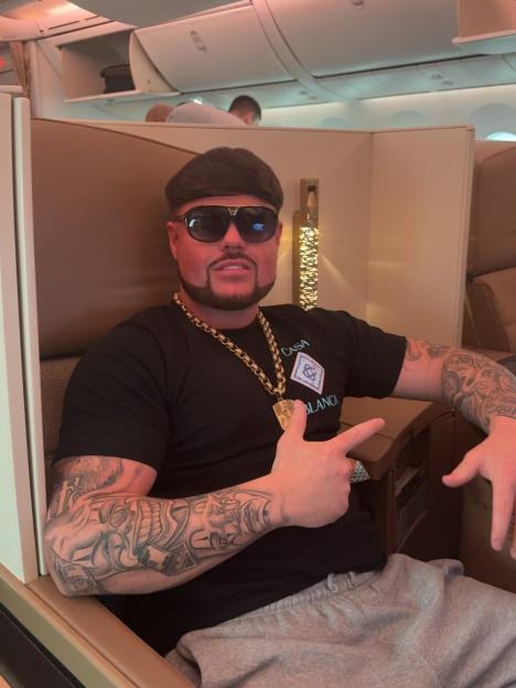 A man with a haircut and beard, wearing sunglasses, a gold chain, and a black t-shirt, sitting on an airplane.