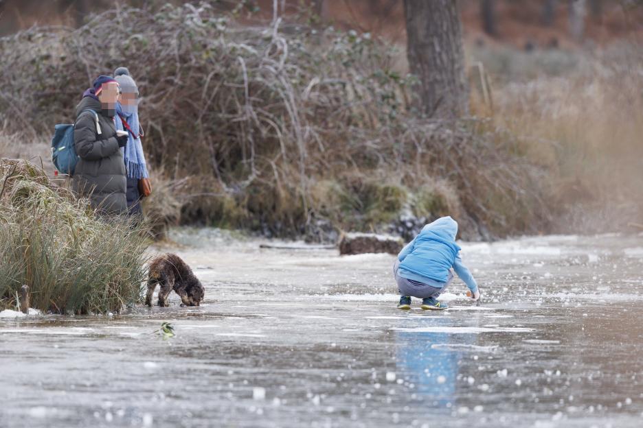 A child crouched on ice hitting it with a hammer as two adults watch from behind.