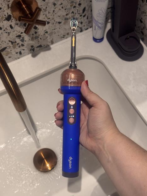 A hand holds a blue and copper Dyson toothbrush over a sink with running water.