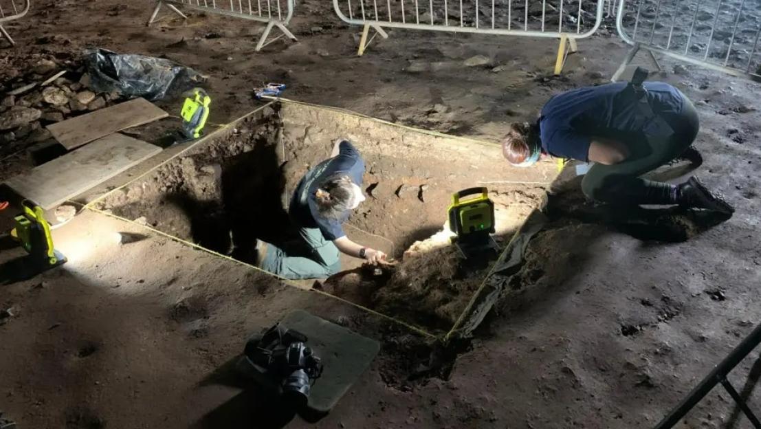 Archaeologists excavating a trench at night, illuminated by portable floodlights.