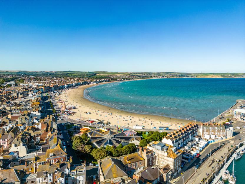 Aerial view of Weymouth Beach and town, with people on the sandy beach and boats docked along the coast.