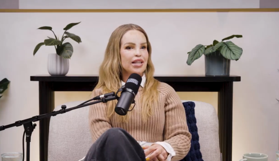 A woman with long blonde hair, wearing a beige sweater and white collared shirt, sits and speaks into a microphone.