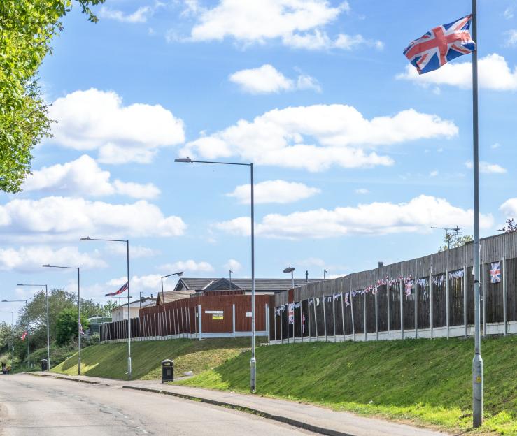 A road next to a grassy hill, with a tall wooden fence adorned with small Union Jack flags, and a large Union Jack flag flying from a flagpole on the right.