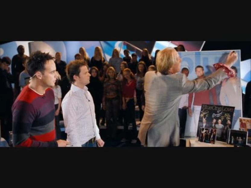 A man in a light suit and a man in a striped shirt look at a poster of a band, while an audience watches in the background.