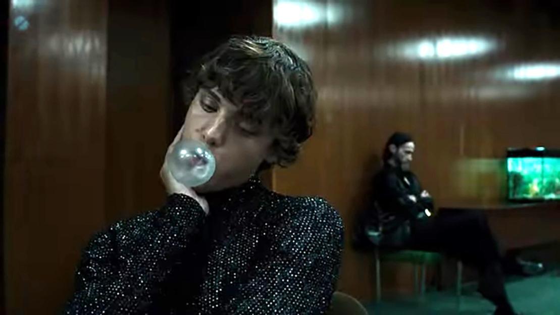 A man blowing a bubble and another man sitting in the background.