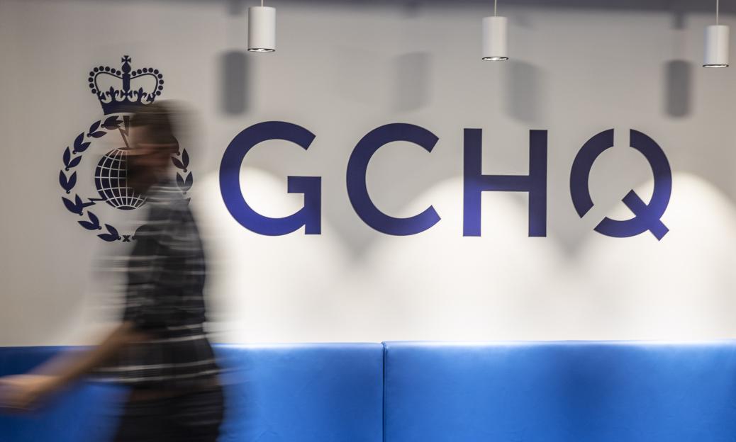 Blurred person walking past a large "GCHQ" sign and logo on a white wall above a blue bench.