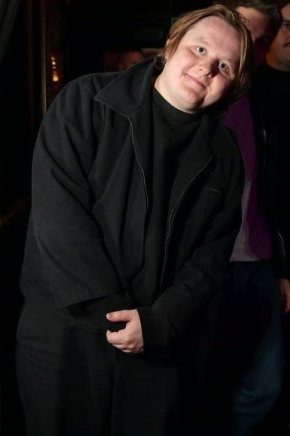 Lewis Capaldi poses in a black jacket and shirt, smiling at the camera.