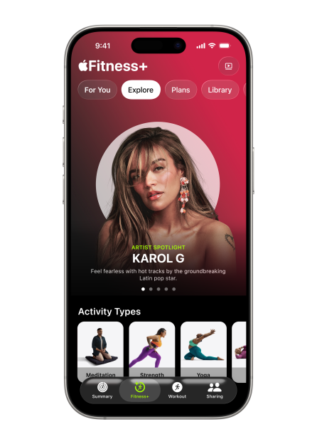 A phone displaying the Apple Fitness+ "Explore" tab, featuring Karol G as the artist spotlight, and showing activity types like Meditation, Strength, and Yoga.