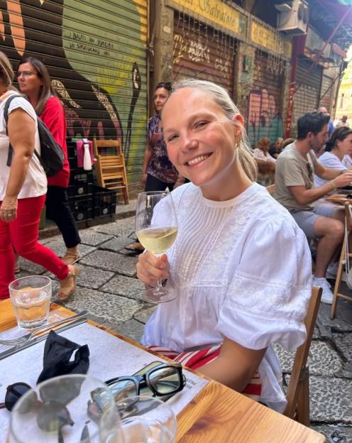 A smiling woman in a white blouse holds a glass of white wine at an outdoor cafe.