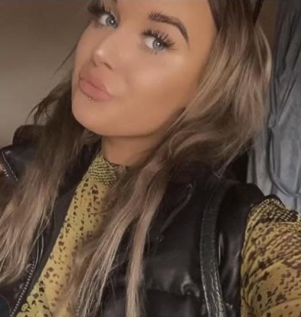 Hannah Metcalfe, a woman with long brown hair, blue eyes, and full lips with a lip piercing, wearing a yellow patterned top and a black vest.