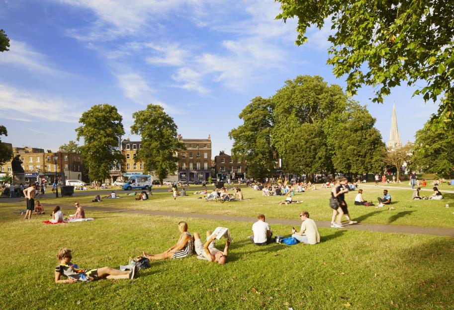 People relaxing in the sun on the grass at Clapham Common, London, with buildings and a church steeple in the background.