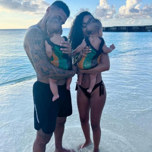A man and a woman stand in the ocean, each holding an infant.