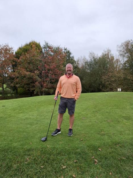 A man in an orange shirt and gray shorts stands on a golf course with a golf club.
