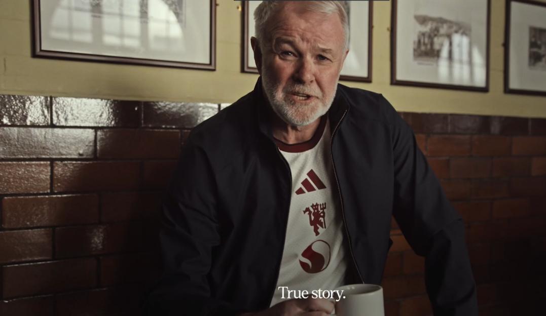A man with a white beard wearing a white t-shirt with red symbols and a black jacket is holding a white cup and talking.