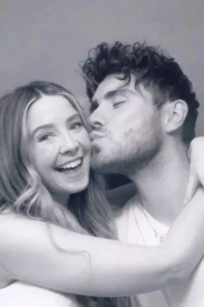 Black and white photo of Zoe Sugg smiling as Alfie Deyes kisses her cheek.