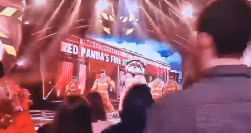 A person in a red panda costume on a stage with others dressed as firefighters, and a large sign that reads "RED PANDA'S FINE ST" in the background.