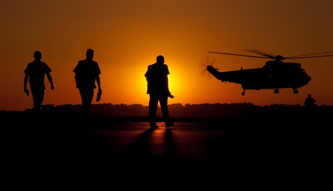 Three men in silhouette walking away from a Royal Navy Sea King helicopter taking off at sunset.