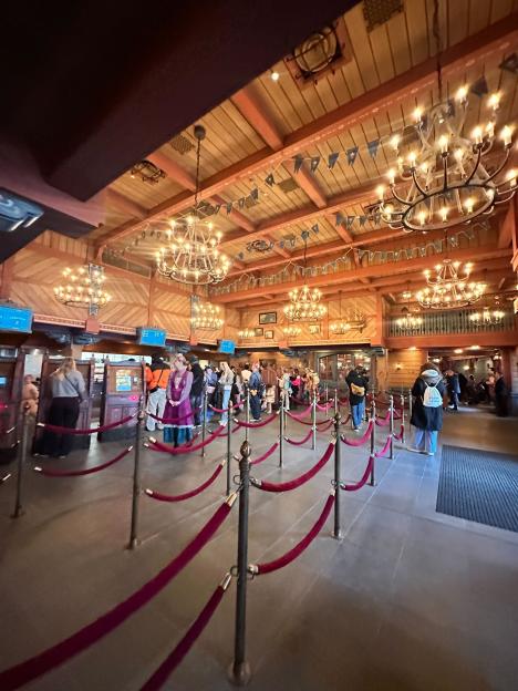 A crowd of people in a large wooden hall with many chandeliers and red velvet ropes.