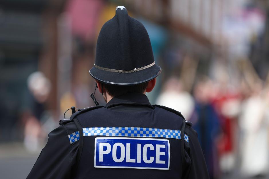 A UK police officer in uniform and helmet, seen from behind.