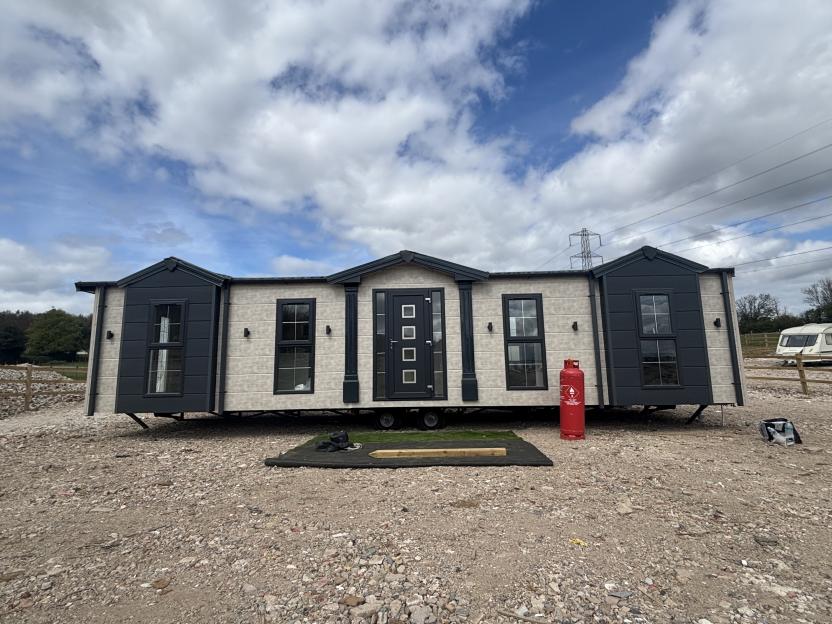 A luxurious mobile home, with dark grey and cream panels and a dark grey front door, stands on a gravel plot under a cloudy sky.