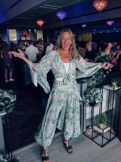 EastEnders star Lorraine Stanley smiles and gestures with her arms wide in a patterned outfit at an event.