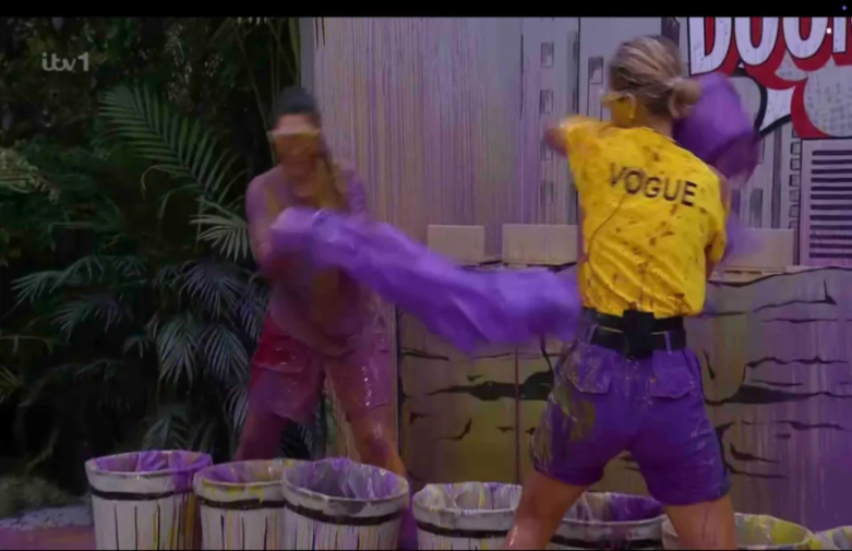 Two people covered in purple liquid, one wearing a yellow "VOGUE" t-shirt, throwing purple items at each other.