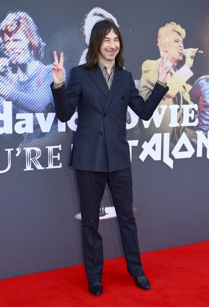 Bobby Gillespie wearing a dark pinstriped suit, black-and-white striped shirt, and black shoes, giving peace signs at the "David Bowie: You're Not Alone" Premiere.