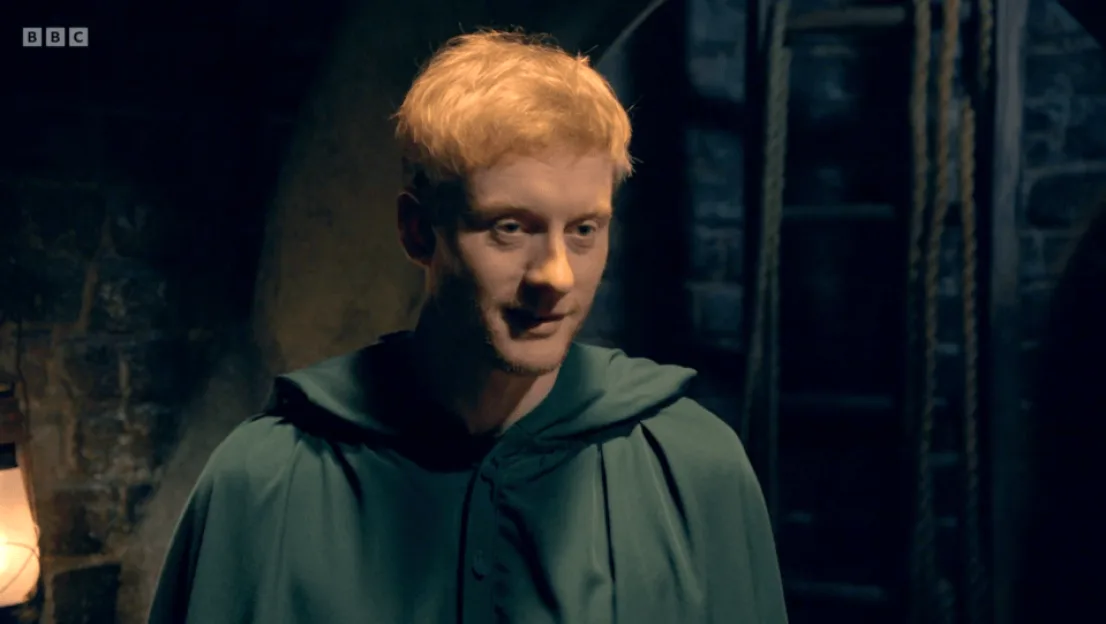 A man with short blonde hair and a green hooded cloak in a dimly lit, rustic setting.