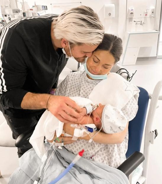 Parents hold their newborn baby in a hospital room, with the baby connected to medical tubes.