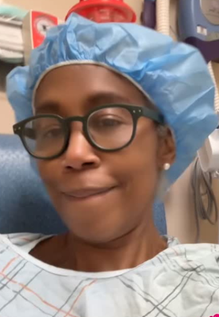 A woman in a hospital gown and surgical cap wearing glasses.