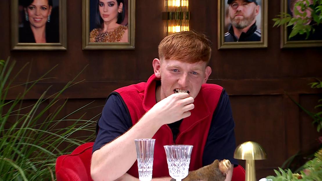 A man wearing a red vest and blue shirt, with red hair and blue eyes, bites into a dish during a trial.