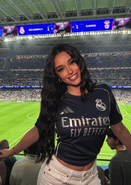 Woman in a Real Madrid jersey smiling at a soccer stadium.