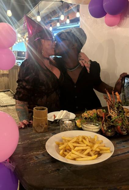 A couple sharing a kiss at a restaurant, surrounded by balloons and food, with the woman wearing a birthday hat.