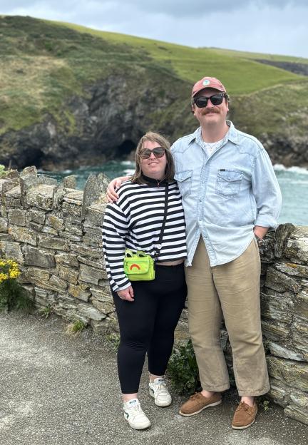 A man and a woman stand next to a stone wall overlooking the ocean and green hills.