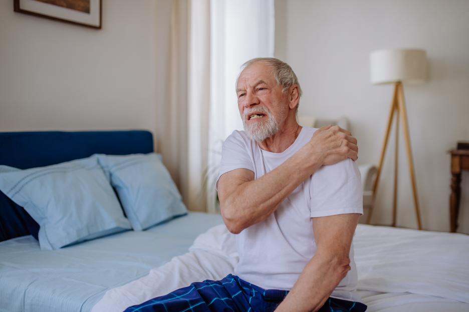 Senior man sitting on a bed and holding his painful shoulder.