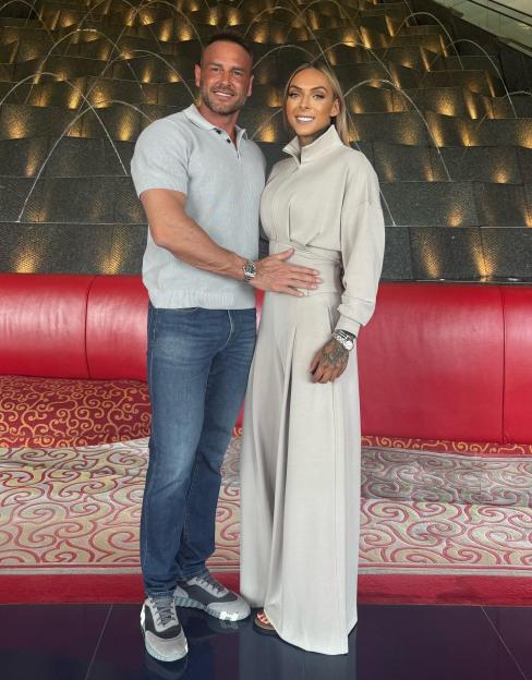 A man and a woman stand smiling, the man with his arm around the woman's waist, in front of a dark, textured wall with water features and golden lighting.