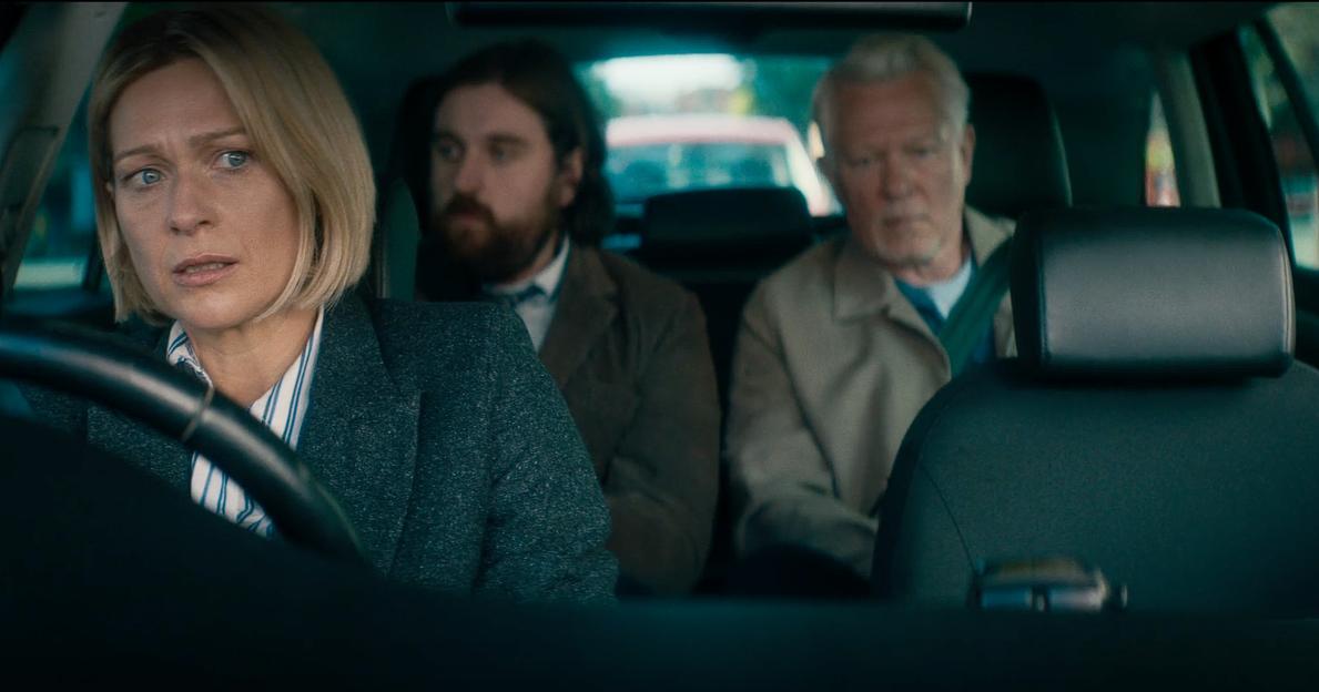 A blonde woman driving a car looks concerned with two men in the backseats.