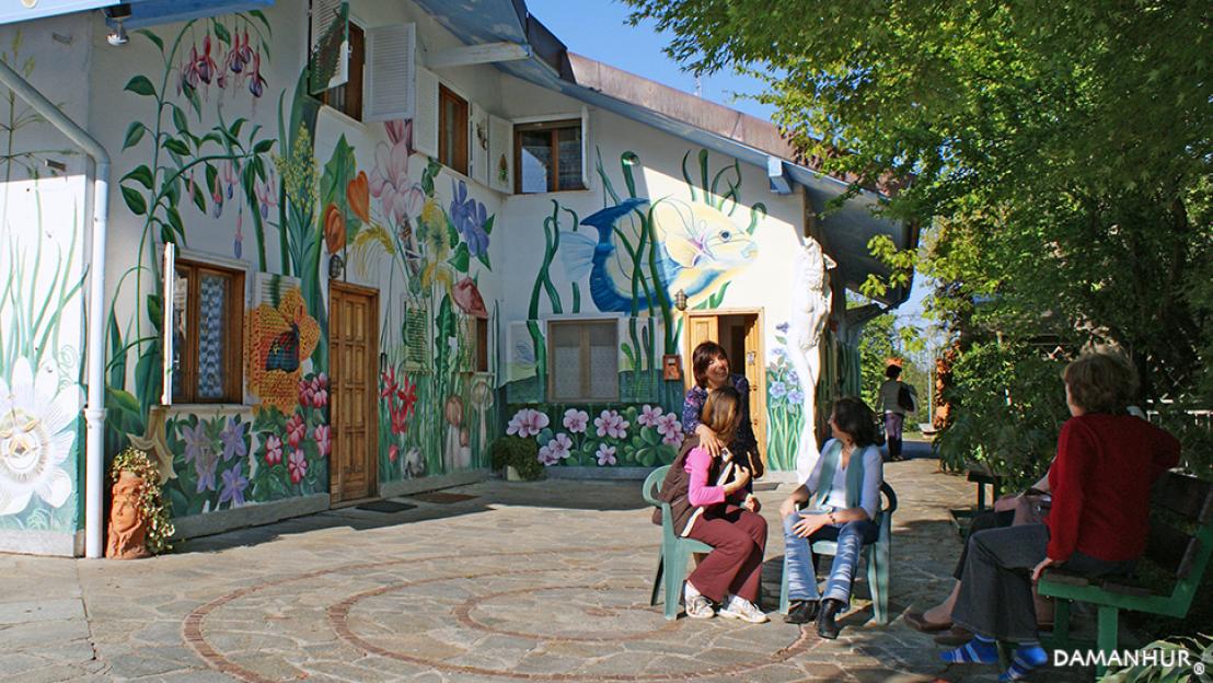 A white house with vibrant murals of flowers, plants, and fish, and several people sitting and talking in a paved courtyard.