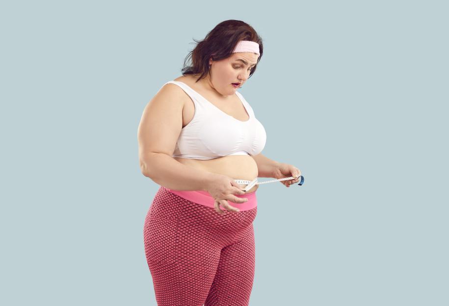 An overweight woman in workout clothes measures her waist with a tape measure, looking shocked at the result.
