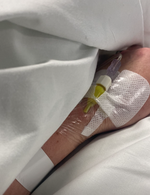 A patient's arm with an IV drip in the hand, bandaged and covered by a white sheet.