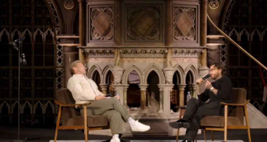 Two men sit in chairs on a stage in front of a carved stone wall.