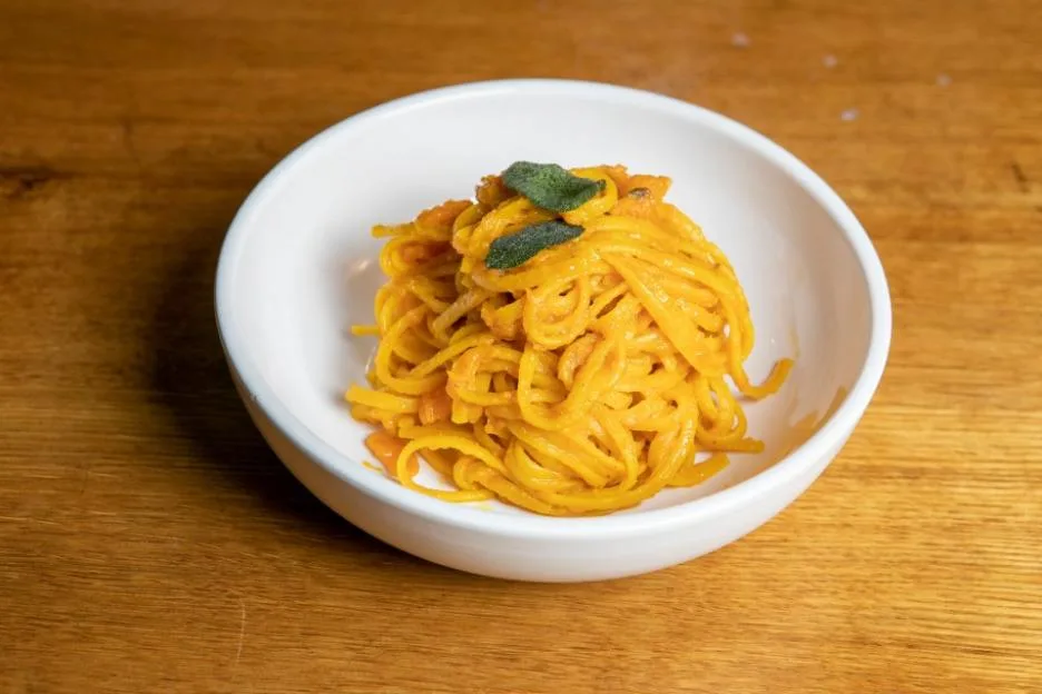 A white bowl filled with pumpkin linguine and topped with sage leaves, set on a wooden table.