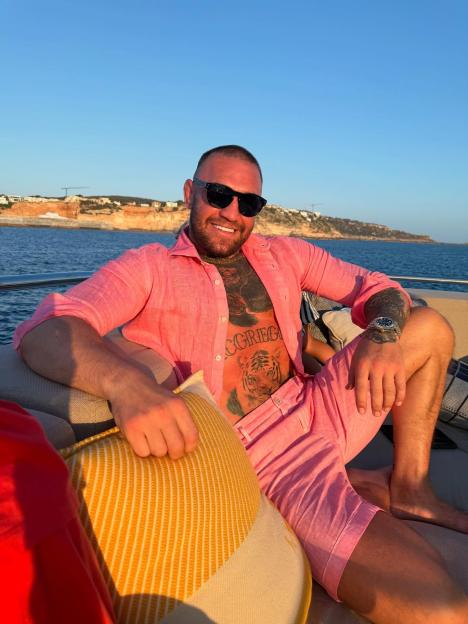 Conor McGregor smiling and relaxing on a boat in a pink shirt and shorts.