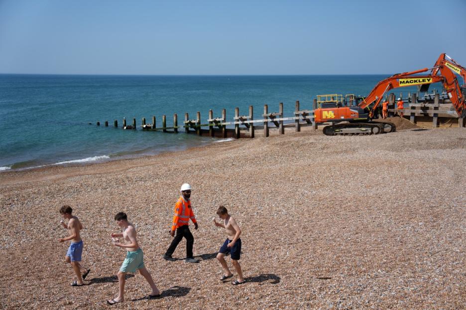Children on a shingle beach walk past an excavator and construction workers building a sea defense.