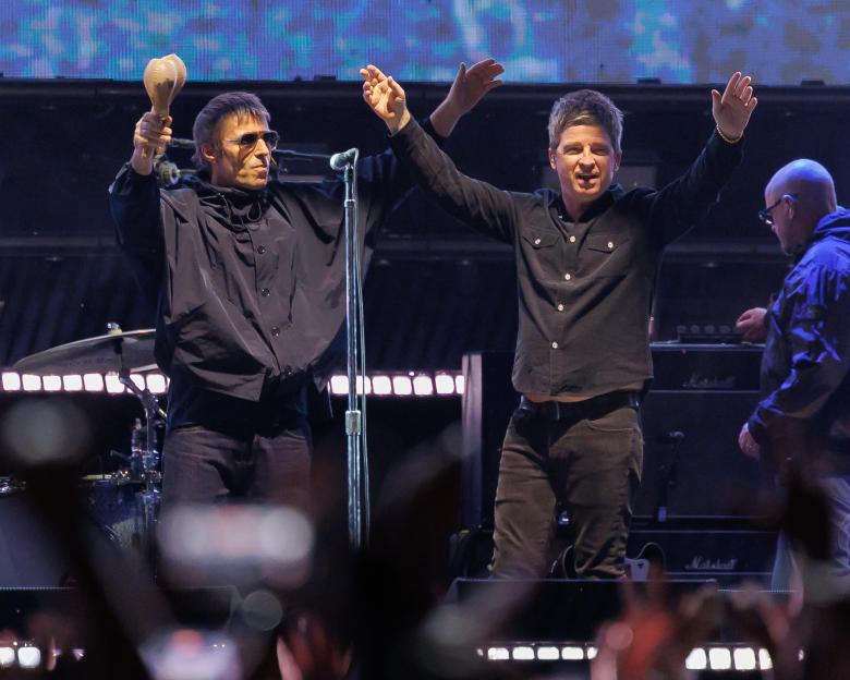 Liam Gallagher holding a maraca and Noel Gallagher with arms raised on stage.